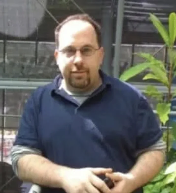 A man, Jay Hartman, standing outdoors near plants, wearing glasses and a navy shirt